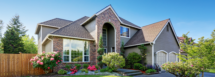 Beige two story brick and siding home with fence and large shrubbery and flowers in front.