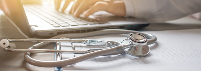 Stethoscope on clipboard with hands typing on laptop in the background.
