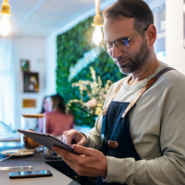 Man wearing apron and holding tablet while standing behind the counter of their bakery business